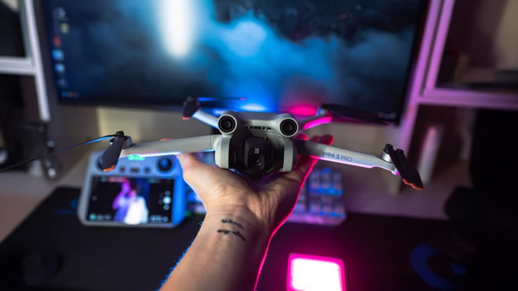 Drone DJI Mini 3 Pro sendo segurado à mão em um ambiente de criador de conteúdo, com iluminação colorida e tela ao fundo.