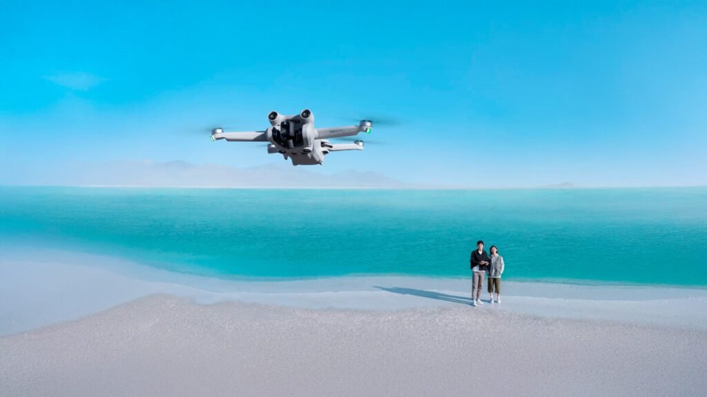 Drone DJI Mini 3 Pro em voo sobre uma praia com mar azul ao fundo, sendo pilotado por um casal.